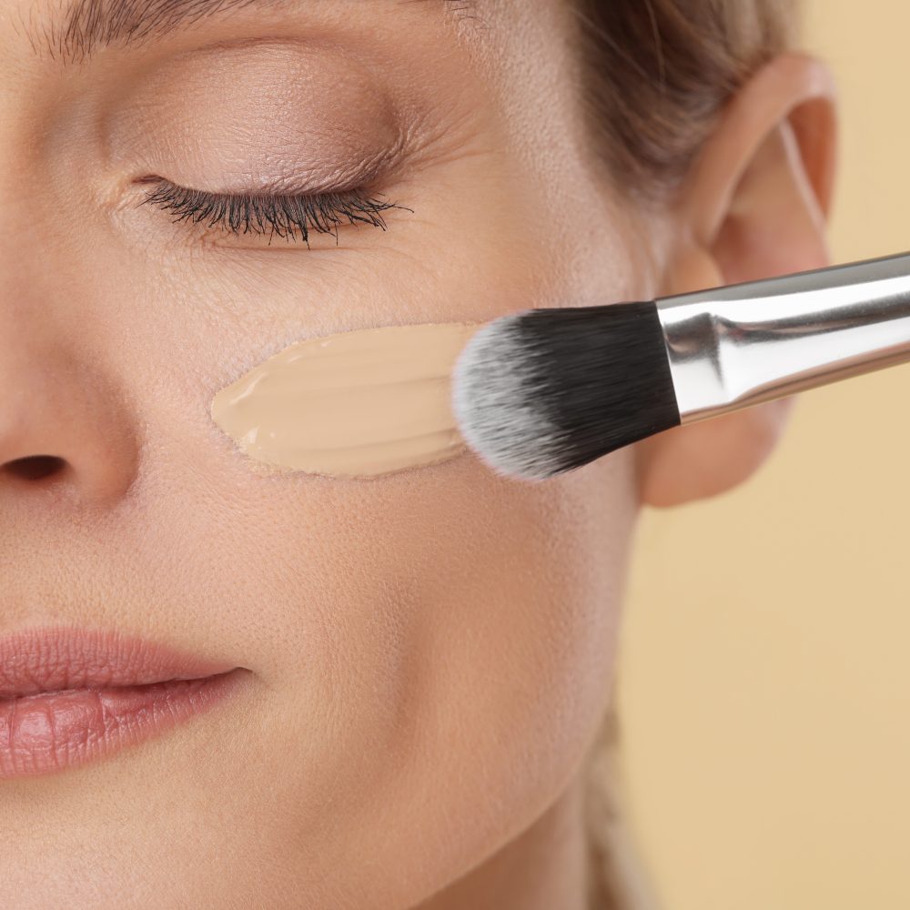 Woman applying foundation on face with brush against beige background, closeup Woman applying foundation on face with brush against beige background, closeup